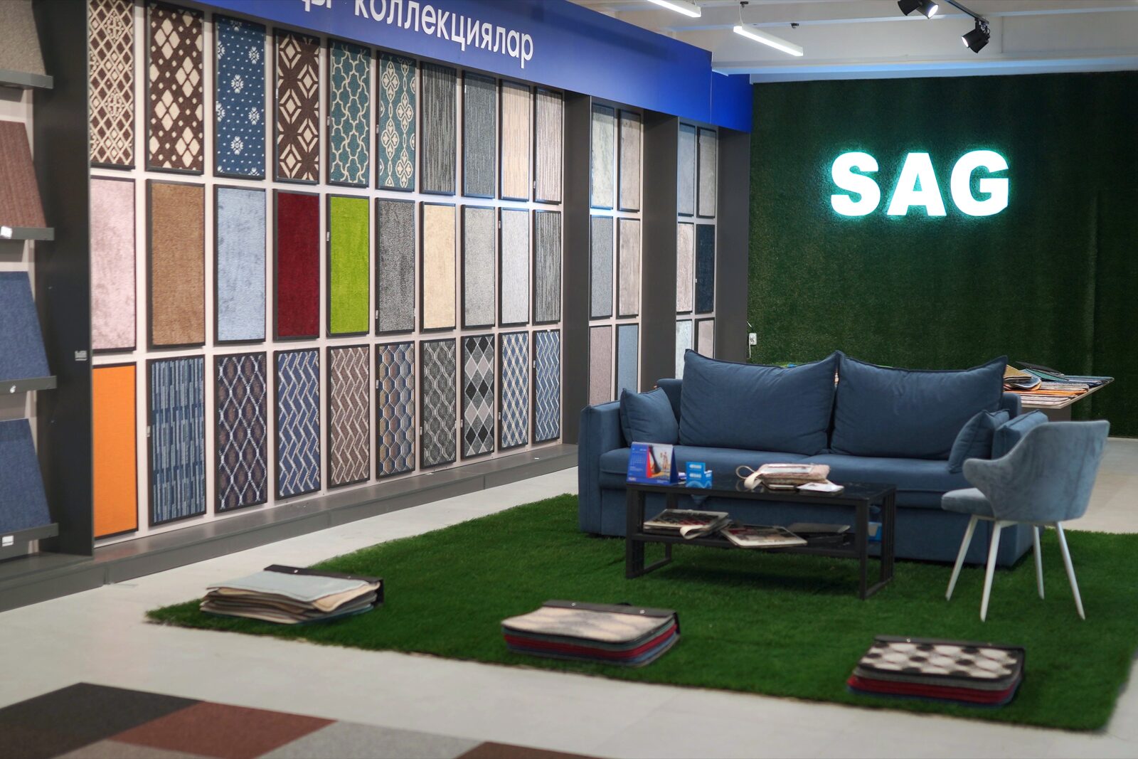 Шоурум SAG Flooring в Бишкеке: стена с образцами ковролина, синий диван на искусственной траве и светящаяся вывеска SAG